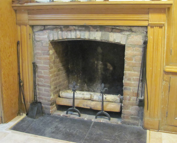 Red brick hearth, encased in a wooden wall. Cemter: wood logs. At the left against the wall, tools for the fire and fireplace maintenance.