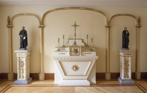 White and gilded Altar table. At the center: a small tabernacle topped by a cross and four chandelier. Two Jesuits statues stand on pillars on both side.