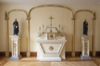 White and gilded Altar table. At the center: a small tabernacle topped by a cross and four chandelier. Two Jesuits statues stand on pillars on both side.