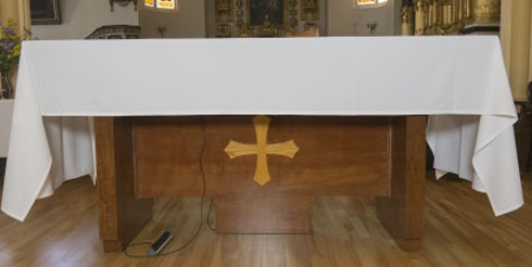 Table autel, rectangulaire en bois. Croix en bois clair appliquée au-devant. Recouverte d’une nappe blanche.