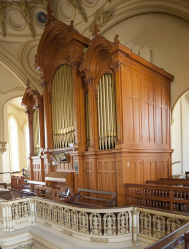 At the balcony: organ side view. Three carved wooden part with a higher central part. Vertical pipes. Front: keyboard and bench. In the foreground, worked railing.