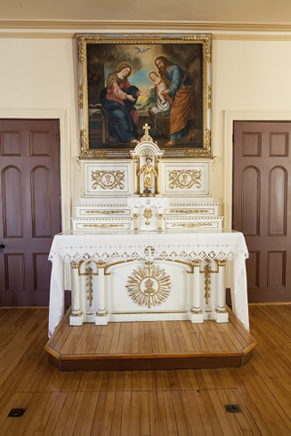 Centered between two doors. Sculpt white and gilded altar and altarpiece. A painting of the Holy Family. Front: Child jesus statuette. Crowned and gilded.