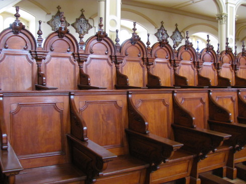 Deux rangées de sièges de bois ouvragé. Plus de quarante sièges. Rangée du fond plus haute avec dossiers en arc et fuseaux décoratifs au haut des dossiers.