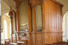 At the balcony: organ side view. Three carved wooden part with a higher central part. Vertical pipes. Front: keyboard and bench. In the foreground, worked railing.