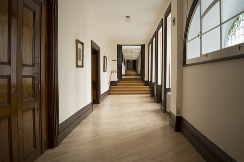Long corridor au plancher de bois entrecoupé par des escaliers de quelques marches. Plusieurs grandes fenêtres sur le mur blanc de droite.