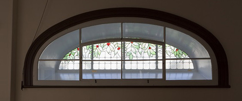 Arched stained-glass window. Located at the top of a wall. Green leaves and red berries on tiles surrounded by tin. At foreground, arched double window with 8 tiles.