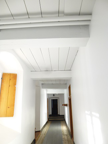 Long white corridor. Wooden door on the left. Large slate floor covered by a long grey carpet. At the middle, the ceiling become vaulted and the floor in stone.