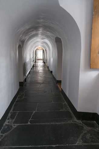 Corridor blanc vouté. Grande fenêtre lumineuse au fond. Murs très épais. Des portes de chaque côté. Plancher de pierres foncées.