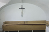 Long wooden container. White room with vaulted ceiling. Large crucifix in the center of the wall.