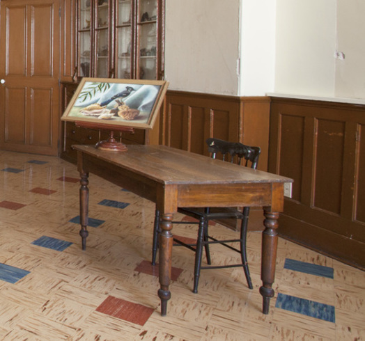 Bureau d’enseignante. Table de bois rectangulaire avec chaise à dossier. Dessus, une peinture didactique de geai bleu dans un arbre.