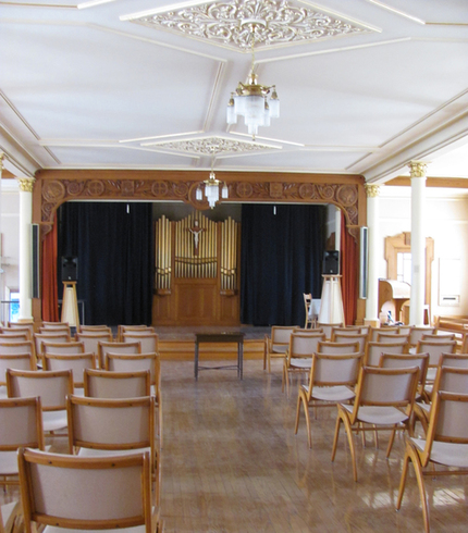 Driveway with chairs on each side. Leading to a scene with dark curtains. Top, a small golden organ. White ceilings, ornaments and crystal chandeliers.