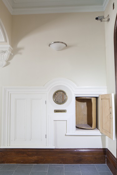 Distinct section of the wall. White door at left. Center: round grid with holes and a mail slot under it. At right, small open door leaving to see a wooden cylinder with openings.