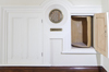 White wall, molding on the floor in dark wood. Circular mesh in the center. Small open door to the left is an open wooden cylinder.