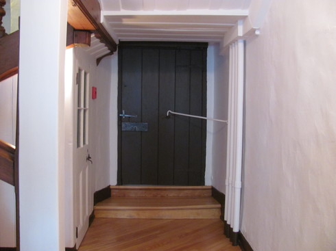 End of corridor. Door in wooden planks painted dark brown. Latch and hand wrought iron. A long iron hook is attached, from the wall to the door, to keep it closed.