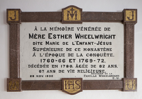 Plaque fixée au mur. Hommage à Mère Esther Wheelwright. Texte en lettres noires sur fond de marbre blanc. Contour brun avec lettres sur fond de dorure.