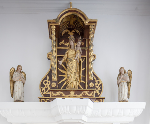 Brown and gilded alcove. Narrow. Reaching the ceiling. Center: Crowned with golden clothes, Virgin Mary with child Jesus in her arms. Both sides: white and golden angel praying.