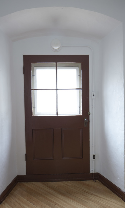 Simple single brown door. Glass window at it center, 4 tiles. Surrounded by white walls.