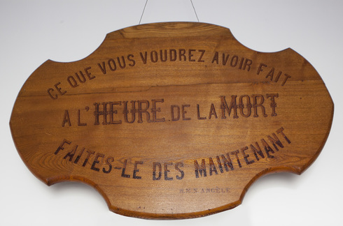 Plaque de bois découpés sur mur blanc. Inscription en trois parties formant un ovale : « Ce que vous voudrez avoir fait à l’heure de votre mort faites le maintenant » N.M.S. Angèle