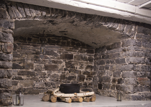 Large fireplace made of grey stones. A the center, wooden logs holding a large black iron-cast cauldron. Two lantern on both sides on the floor.