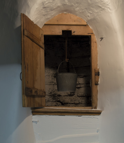 In a thick with wall, a small light wooden door is open. We see the stone wall of the well and a hanging bucket.