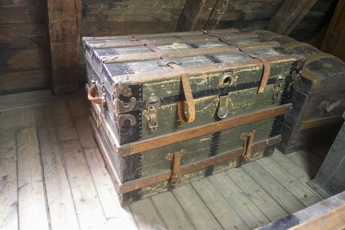 Large coffre rectangulaire en bois peint vert et noir. Des lanières de cuir le maintiennent fermé et servent d’attaches et de ganses sur les côtés.