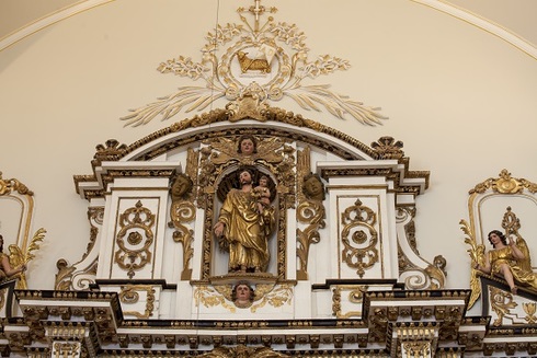 Partie supérieure de l’ornementation du mur de la chapelle. Alcôve blanc et doré surmonté d’un bas-relief d’un Sacré-Cœur. Au centre, Sculpture de St-Joseph tenant l’enfant Jésus dans ses bras.