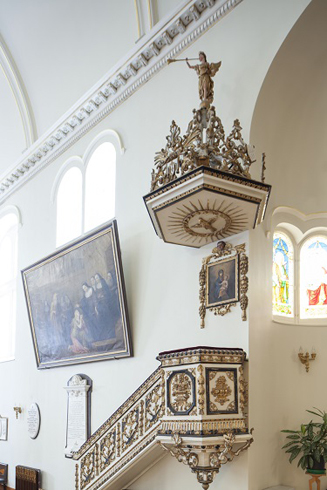 Part of a wall.Hanging carved and gilded Sounding board. An angel  above it holding on his foot. Under, Pulpit looking like a balcony accessed by a stair.