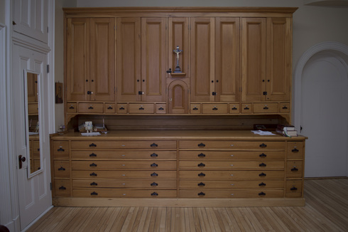 Grand meuble de rangement en bois. Haut : rangée de petits tiroirs et armoires. Au centre : un crucifix. Bas : comptoir et tiroirs. De chaque côté du meuble : des portes blanches.