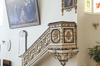 Small circular balcony decorated with white and gold. Upper area, golden and gilded, overhung by an angel with a trumpet, standing on the point of a foot.