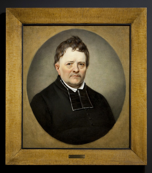 Simple wood frame. Painted portrait in an oval. Abbot Maguire in his chaplain clothes. Shaved, brown messy  hair, brown eyes. Looking to the left.