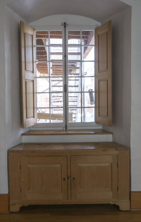Sous une fenêtre à carreaux avec volets de bois, un coffre de bois clair construit pour s’encastrer dans l’espace précis sous la fenêtre.