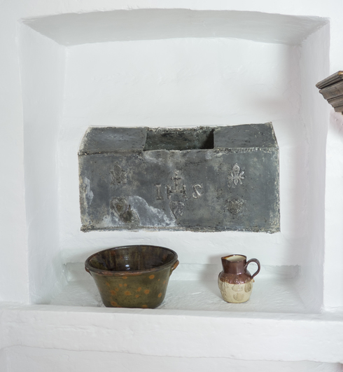 In a white alcove, encased iron box. Opening at the center, on top. Hammered and embossed with the letters HIS. Bowl and small jug on a shelf.