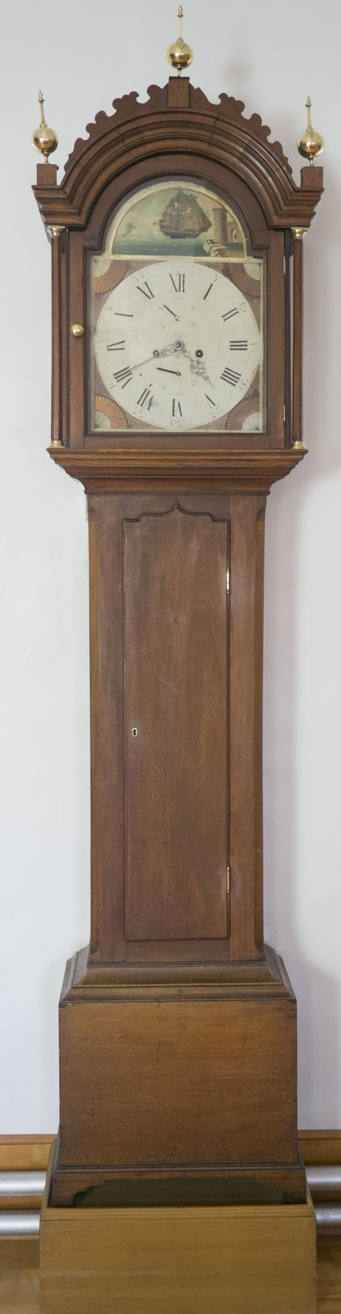 Brown wood Grandfather clock. Roman numeral. Up the dial, a ship. Arched top finish, carved and ornated with three gilded spindle.