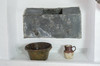 In a white alcove, encased iron box. Opening at the center, on top. Hammered and embossed with the letters HIS. Bowl and small jug on a shelf.